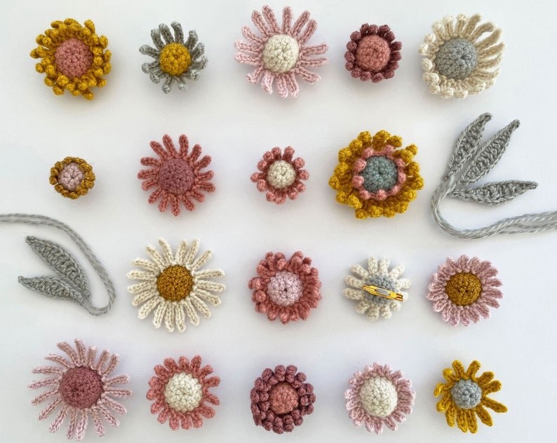 May include: A collection of 17 crocheted flower pins in various shades of pink, yellow, and gray. The flowers are all different sizes and shapes, and some have a center button.