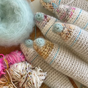 K&ouml;nnte beinhalten: Nahaufnahme von mehreren geh&auml;kelten Garnkreationen in verschiedenen Beige-, Wei&szlig;- und Blaut&ouml;nen. Das Garn ist weich und flauschig, und die geh&auml;kelten St&uuml;cke sind filigran und detailliert. Im Hintergrund befinden sich auch mehrere Garnkn&auml;uel, darunter ein hellblaues Garnkn&auml;uel.