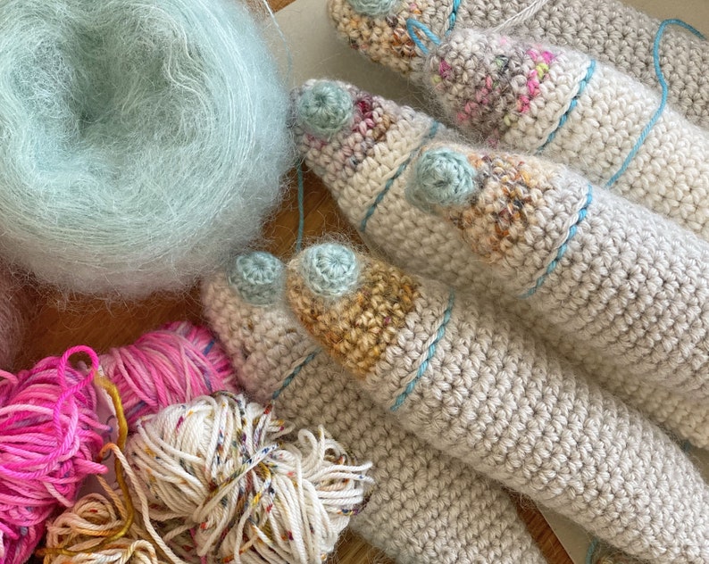 K&ouml;nnte beinhalten: Nahaufnahme von mehreren geh&auml;kelten Garnkreationen in verschiedenen Beige-, Wei&szlig;- und Blaut&ouml;nen. Das Garn ist weich und flauschig, und die geh&auml;kelten St&uuml;cke sind filigran und detailliert. Im Hintergrund befinden sich auch mehrere Garnkn&auml;uel, darunter ein hellblaues Garnkn&auml;uel.
