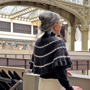 May include: A dark gray and white knitted cape with a ruffled edge. The cape features horizontal stripes and is worn over a black turtleneck. The person is wearing cream-colored pants and is standing in a building with a glass ceiling.