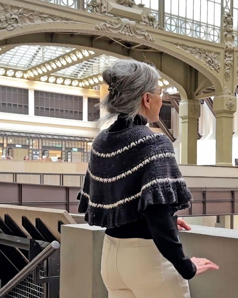 May include: A dark gray and white knitted cape with a ruffled edge. The cape features horizontal stripes and is worn over a black turtleneck. The person is wearing cream-colored pants and is standing in a building with a glass ceiling.