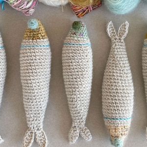 K&ouml;nnte beinhalten: Sechs geh&auml;kelte Fischspielzeuge in hellbeige mit blauen und gelben Akzenten. Die Fische sind in einer Reihe auf einer hellgrauen Oberfl&auml;che angeordnet.
