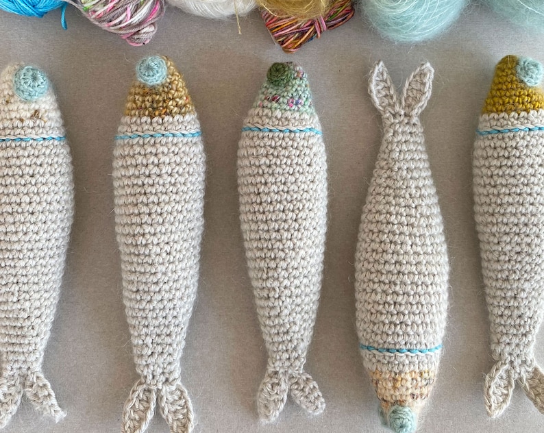 K&ouml;nnte beinhalten: Sechs geh&auml;kelte Fischspielzeuge in hellbeige mit blauen und gelben Akzenten. Die Fische sind in einer Reihe auf einer hellgrauen Oberfl&auml;che angeordnet.