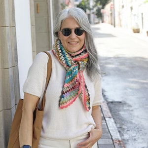 May include: A woman with gray hair is wearing a white sweater and a colorful crocheted scarf. She is wearing sunglasses and has a brown leather bag over her shoulder.