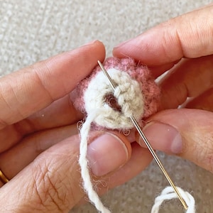 May include: A close-up of a hand holding a small, crocheted, pink and white ball with a needle and white thread going through it.