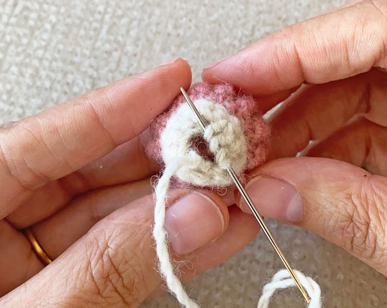 May include: A close-up of a hand holding a small, crocheted, pink and white ball with a needle and white thread going through it.