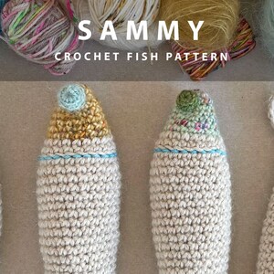 K&ouml;nnte beinhalten: Vier geh&auml;kelte Fisch-Amigurumi-Spielzeuge in verschiedenen Farben. Die Fische sind aus wei&szlig;em Garn gefertigt und haben unterschiedlich farbige Details an ihren K&ouml;pfen. Die Fische sind in einer Reihe auf einer braunen Oberfl&auml;che angeordnet. Der Text "SAMMY CROCHET FISH PATTERN" ist &uuml;ber den Fischen angezeigt.