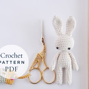 May include: A crochet pattern PDF for making a white amigurumi bunny. The pattern is shown next to a pair of gold scissors and a wooden ruler.