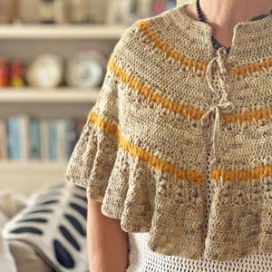 May include: A beige crochet cape with horizontal yellow stripes. The cape has a ruffled hem and a tie closure at the neckline. The garment is worn over a white, open-weave top.