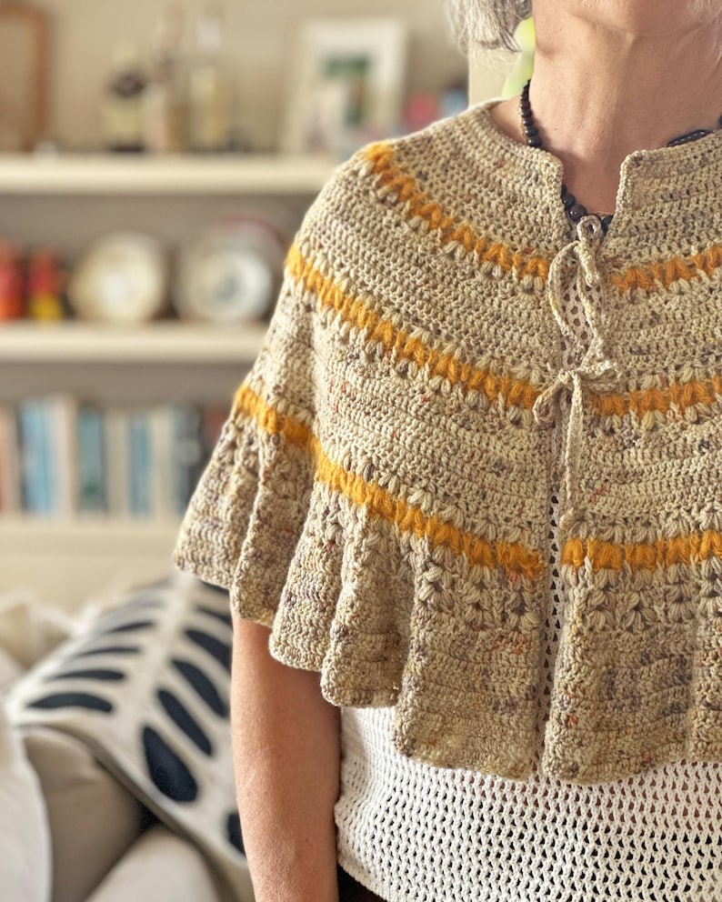 May include: A beige crochet cape with horizontal yellow stripes. The cape has a ruffled hem and a tie closure at the neckline. The garment is worn over a white, open-weave top.