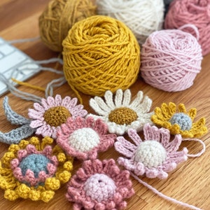 May include: A collection of crocheted flowers in various colors, including pink, yellow, and gray. The flowers are arranged on a wooden surface with balls of yarn in the background.