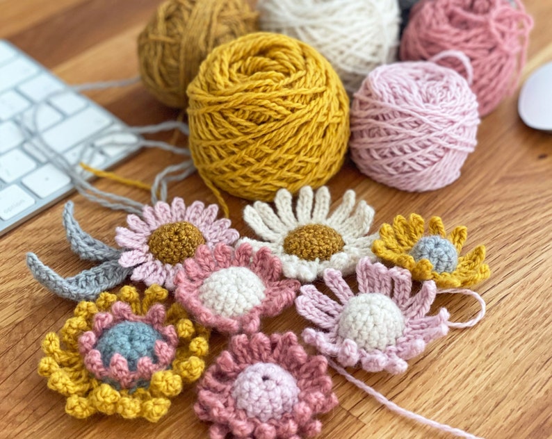 May include: A collection of crocheted flowers in various colors, including pink, yellow, and gray. The flowers are arranged on a wooden surface with balls of yarn in the background.