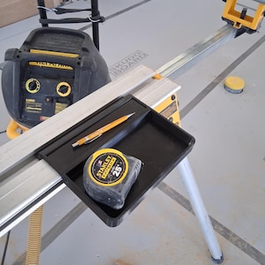 May include: A black plastic tool tray attached to a metal miter saw stand, containing a yellow pencil and a Stanley FatMax 25-foot tape measure. A black and yellow DeWalt air compressor sits in the background near the metal stand rails.