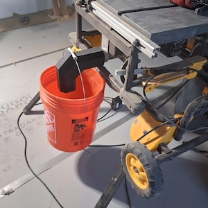 May include: A black 3D-printed dust collection attachment mounted to a table saw, positioned to direct debris into a bright orange plastic bucket. The bucket features a white logo and warning label on its side, with a metal handle.