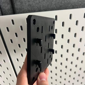 Pode incluir: Uma placa de montagem quadrada de plástico preto com múltiplos orifícios circulares e vários ganchos em forma de L salientes na sua superfície. A placa tem um acabamento texturado e foi concebida para ser fixada num painel perfurado branco.