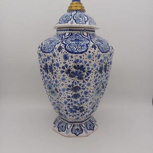 Op de afbeelding: Een witte keramische gemberpot met een afneembaar deksel, voorzien van ingewikkelde blauwe bloem- en krulpatronen over het lichaam en het deksel. Het deksel is bekroond met een kleine, goudkleurige knop en de voet heeft een zeshoekige vorm.
