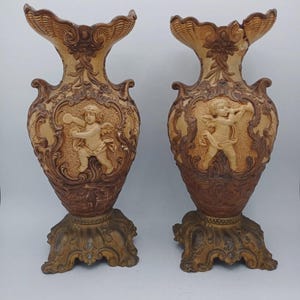 May include: A pair of tall, ornate vases featuring a two-tone brown and cream finish. Each vase displays a central relief of a cherub figure surrounded by intricate scrollwork and floral motifs, set upon a wide, decorative pedestal base.