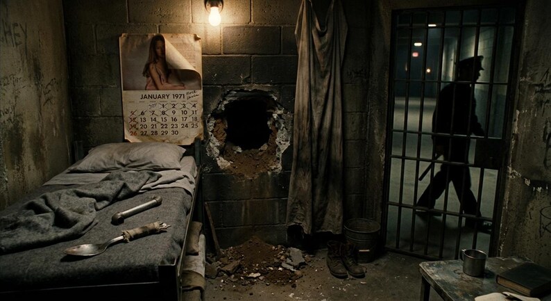 May include: A prison cell interior featuring a metal bed frame with a gray blanket, a large metal spoon, and a pipe. A January 1971 calendar hangs on the cinder block wall next to a large hole. A metal door with bars stands to the right.