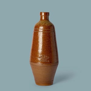 Puede incluir: Una botella de gres alta y cónica con un esmalte marrón brillante y moteado en la sección superior y una base marrón mate y terrosa. La botella presenta un cuello estrecho, un borde pequeño y un logotipo de corona en relieve con el texto PORTO.