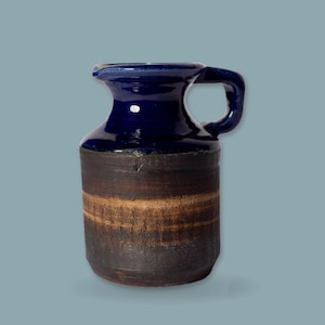 Può includere: Una brocca in ceramica con una sezione superiore smaltata in blu scuro lucido e un corpo inferiore testurizzato in marrone scuro opaco. Il recipiente presenta un bordo svasato, un manico laterale singolo e una forma cilindrica con una fascia orizzontale marrone chiaro.