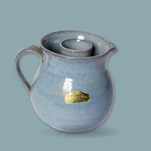 Peut inclure: Une cruche en céramique bleu clair avec un corps arrondi, une anse incurvée et un insert cylindrique central. Un autocollant rectangulaire doré sur le devant porte le texte GRES LE REFUGE en lettres noires, avec du texte plus petit en dessous.