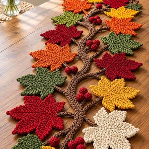 Pode incluir: Um caminho de mesa em crochê com uma estrutura central de ramos castanhos, cachos de pequenas bagas vermelhas e várias folhas de ácer em tons de vermelho, laranja, amarelo, verde e creme, dispostas num padrão decorativo ao longo do comprimento.