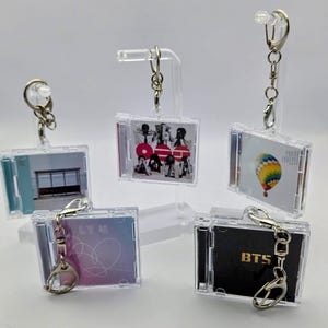 May include: Five miniature clear plastic CD case keychains featuring various album cover art. One shows a bus stop, another a group of people with red circles, one a hot air balloon, one a pink and blue heart, and one with the text BTS.