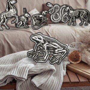 May include: Black and white animal skeleton cutouts, including a horse, rabbit, rat, snake, cat, and frog. The cutouts are arranged on a bed and a wooden table with a book and dried flowers. The frog cutout is on a gray knitted blanket.
