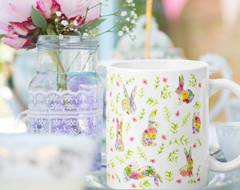 Könnte beinhalten: Weiße Keramiktasse mit einem sich wiederholenden Muster aus bunten Blumenhasen und grünen Blättern. Die Tasse hat einen gebogenen Henkel. Der Hintergrund zeigt eine Vase mit Blumen. Ein Produkt zum Thema Frühling oder Ostern.