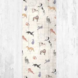 A beige modal scarf with illustrated greyhound dog pattern on it laid out on a grey wooden table.