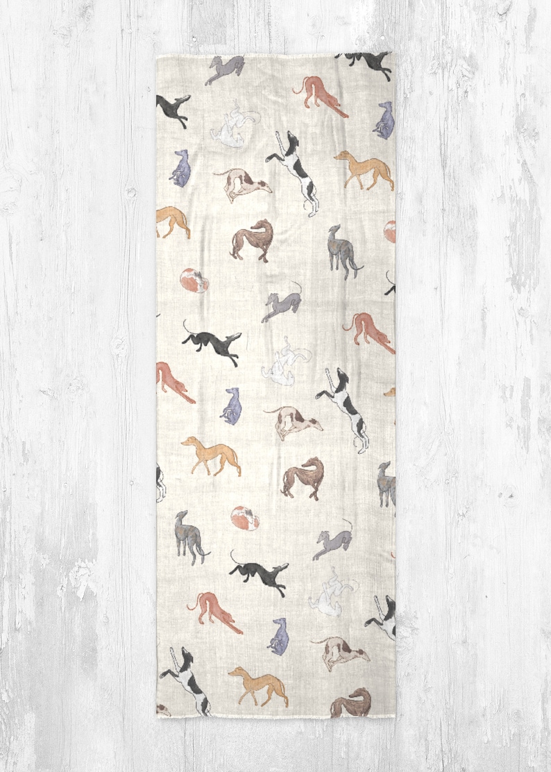 A beige modal scarf with illustrated greyhound dog pattern on it laid out on a grey wooden table.