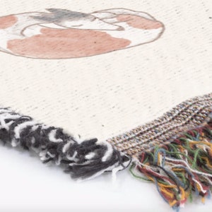 K&ouml;nnte beinhalten: Cremefarbene gewebte Decke mit einer Illustration eines schlafenden Fuchses in Braun- und Graut&ouml;nen. Die Decke hat einen Fransenrand mit schwarz-wei&szlig;en Quasten auf einer Seite und einer mehrfarbigen Franse auf der anderen.