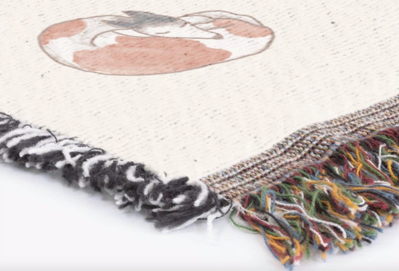 K&ouml;nnte beinhalten: Cremefarbene gewebte Decke mit einer Illustration eines schlafenden Fuchses in Braun- und Graut&ouml;nen. Die Decke hat einen Fransenrand mit schwarz-wei&szlig;en Quasten auf einer Seite und einer mehrfarbigen Franse auf der anderen.
