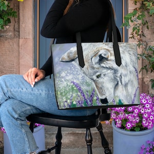 Può includere: Una borsa tote nera con un'illustrazione di un lupo bianco e fiori viola. Il lupo guarda a sinistra e ha uno sfondo bianco con un motivo floreale verde chiaro e viola.