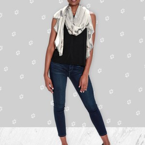 A woman wearing around her neck a beige modal scarf with illustrated greyhound dog pattern with black tank top, jeans, and black heels.