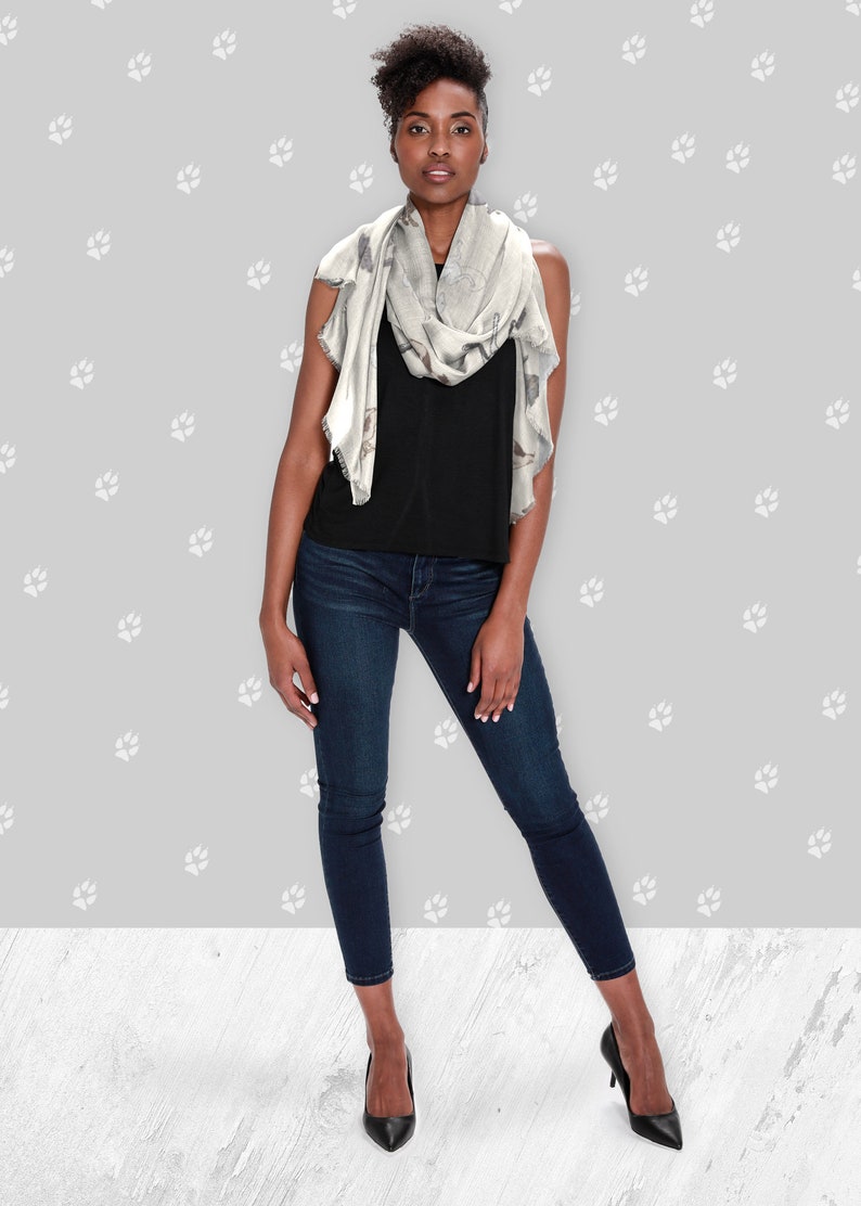 A woman wearing around her neck a beige modal scarf with illustrated greyhound dog pattern with black tank top, jeans, and black heels.