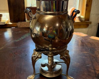 Op de afbeelding: Een zilverkleurige metalen samovar met een bolvormig lichaam, een tapkraan met een zwarte hendel en twee zijhandgrepen. Het vat rust op drie sierlijke, gebogen poten en heeft een afneembaar deksel met een doorzichtige glazen knop.
