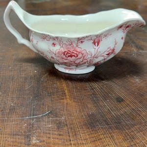 May include: A cream-colored ceramic gravy boat featuring a delicate red floral pattern. The vessel has a curved handle, a wide pouring spout, and a scalloped rim, decorated with detailed illustrations of blooming flowers and buds.