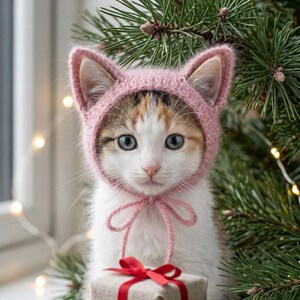 May include: A calico kitten with white, brown, and black fur wearing a soft, light pink knitted bonnet with pointed cat ears. The bonnet features thin knitted ties tied in a bow under the chin. A small gift box with a red ribbon is visible.