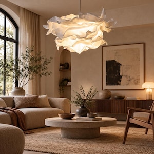 Pode incluir: Uma grande luminária pendente branca em forma de nuvem, com múltiplos painéis de tecido translúcido e franzido que emitem um brilho quente, suspensa do teto por uma haste metálica fina e reta em uma sala de estar.