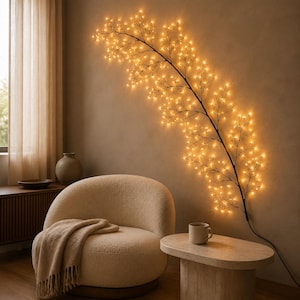Op de afbeelding: Een decoratieve taklamp voor aan de muur met een donkere, slanke steel versierd met talloze kleine, warm gloeiende LED-lampjes. De tak strekt zich diagonaal over de muur uit en verspreidt een zachte, gouden verlichting die de vorm benadrukt.