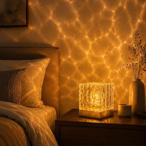 Könnte beinhalten: Eine quadratische Tischleuchte mit einem strukturierten, durchscheinenden Glasschirm, der ein warmes, goldenes Licht ausstrahlt. Die Lampe hat einen Metallsockel mit einem sichtbaren Einschaltknopf und wirft ein wellenförmiges Muster an die Wand.