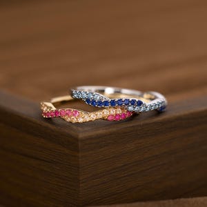 May include: Two intertwined rings featuring a wavy design. One ring is crafted from silver-toned metal and set with blue stones. The second ring is crafted from gold-toned metal and set with pink and clear stones along the wavy band surface.