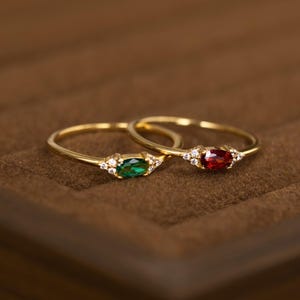 May include: Two thin gold rings, one featuring an oval-cut green gemstone and the other an oval-cut red gemstone. Each ring is accented with three small clear stones on either side of the central oval gem, set on a slender gold band.