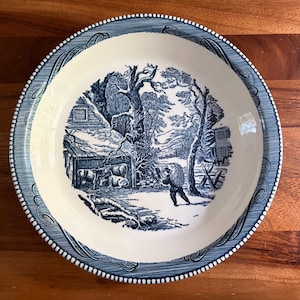 May include: A round ceramic plate with a cream-colored center featuring a blue transferware illustration of a winter farm scene with cattle, a barn, and a person walking. The rim is decorated with a blue and white patterned border design.