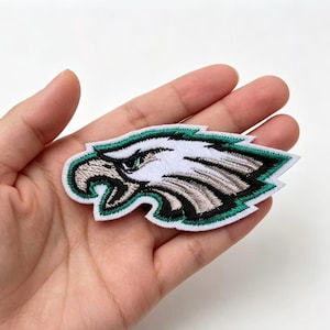 May include: An embroidered patch featuring a stylized eagle head in profile. The design uses white, black, grey, and teal thread colors with a white border. The eagle is depicted with a sharp beak, a focused eye, and layered feathers in a horizontal orientation.