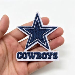 Puede incluir: Un parche de tela bordado que presenta una estrella azul marino con un borde blanco, colocado sobre una base rectangular blanca con la palabra COWBOYS cosida en letras mayúsculas azul marino. El diseño utiliza una estrella y texto.
