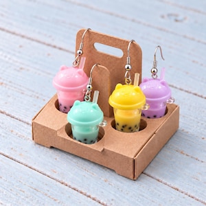 May include: Four dangle earrings shaped like miniature bubble tea cups with cat-ear lids, featuring pink, mint green, yellow, and purple colors with black tapioca pearls inside, all held in a small brown cardboard drink carrier with a handle.