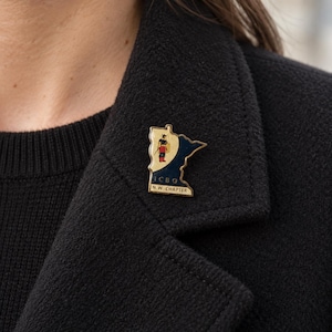 Puede incluir: Un pin de solapa de metal dorado con la forma del estado de Minnesota, con un diseño en color crema y azul oscuro. El pin muestra un pequeño gráfico de una figura con casco y el texto ICBO N.W. CHAPTER impreso en negro en la parte inferior.