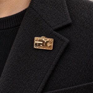 May include: A rectangular gold-toned metal lapel pin featuring the embossed text IRC construction and the number 50. The pin is attached to the lapel of a black textured wool-blend blazer worn over a black knit sweater, showcasing a metallic finish.